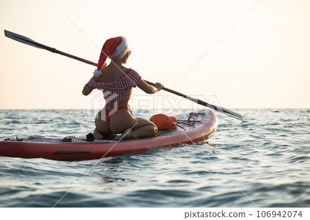 sexy girl in a Santa Claus hat floats on the sea on a Sup Board in a red bikini with a paddle sexy girl in a Santa Claus hat floats on the sea on a Sup Board in a red bikini with a paddle 106942074