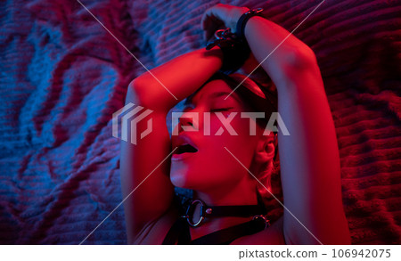 portrait of a sexy girl in a leather mask and bdsm accessories on a bed enjoying sex in orgasm with emotions on her face in neon light portrait of a sexy girl in a leather mask and bdsm accessories on a bed enjoying sex in orgasm with emotions on her face in neon light 106942075