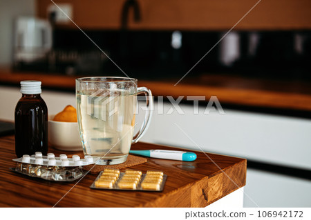 Natural and medical cold and flu remedies on table at home. Cold and flu influenza fall autumn and winter season. Cup with hot tea, lemon, thermometer pills and capsules Natural and medical cold and flu remedies on table at home. Cold and flu influenza fall autumn and winter season. Cup with hot tea, lemon, thermometer pills and capsules 106942172