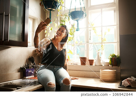 Woman taking a selfie with a phone or video call and smiling in the kitchen at home. Edgy, curvy and relaxed female posting photos on social media app and using online chat in the kitchen Woman taking a selfie with a phone or video call and smiling in the kitchen at home. Edgy, curvy and relaxed female posting photos on social media app and using online chat in the kitchen 106942844