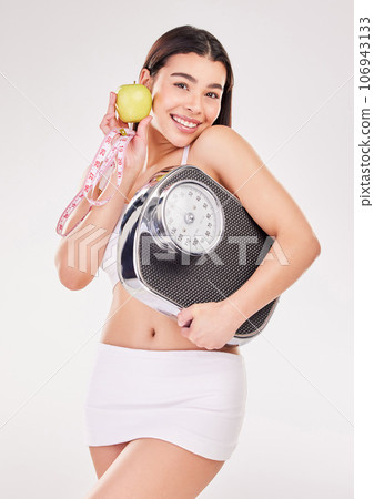 An apple a day.... Studio shot of an attractive young woman posing with an apple, measuring tape and weightscale against a grey background. An apple a day.... Studio shot of an attractive young woman posing with an apple, measuring tape and weightscale against a grey background. 106943133