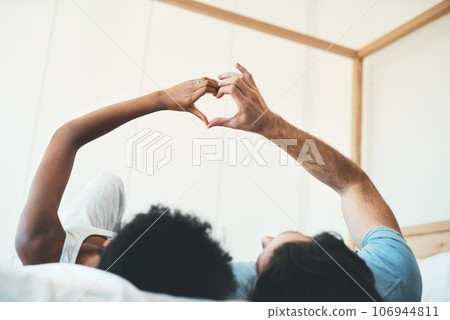 Interracial couple, hands and heart emoji in bed for morning, love or care relaxing together at home. Man and woman lying in bedroom with touching hand for loving symbol, sign or gesture at the house 106944811