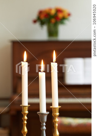 Burning candles in a cosy room against a blurry background. Lighting a candle symbolizes hope and faith in religion. Candle lights in gold and silver holders, for a wedding, funeral home, or church Burning candles in a cosy room against a blurry background. Lighting a candle symbolizes hope and faith in religion. Candle lights in gold and silver holders, for a wedding, funeral home, or church 106945020