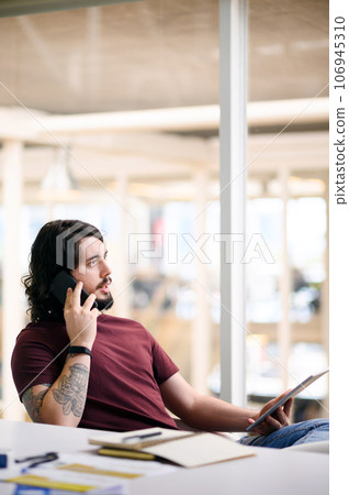 Do you have any news for me. Shot of a young businessman talking on a cellphone while using a digital tablet in an office. Do you have any news for me. Shot of a young businessman talking on a cellphone while using a digital tablet in an office. 106945310