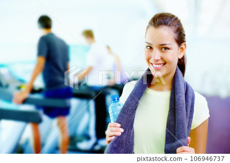 Young woman smiling. Portrait of young smiling woman holding water bottle and towel with people working out in background. 106946737
