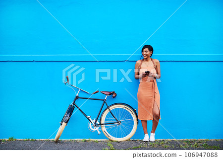 Bicycle, young woman with smartphone and happy outside by blue wall. Cycling in urban area, health wellness and female person on cellphone outdoors standing in streets with bike or cycle in road 106947088