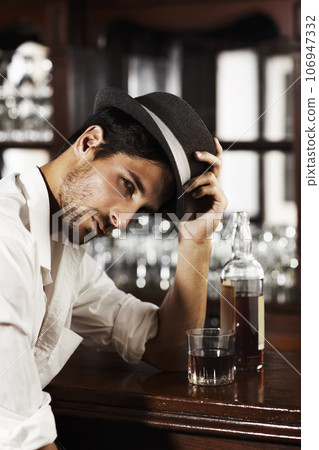 Portrait, whisky and a man sitting at the bar drinking alcohol in a glass while alone or feeling unhappy. Brandy, drink and beverage with a handsome young male pub customer at a wooden counter Portrait, whisky and a man sitting at the bar drinking alcohol in a glass while alone or feeling unhappy. Brandy, drink and beverage with a handsome young male pub customer at a wooden counter 106947332