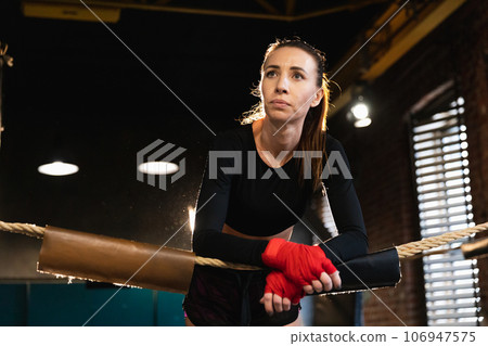 Woman fighter girl power. Woman fighter with red boxing wraps protective bandages standing on boxing ring leaning on ropes waiting resting. Strong powerful girl. Strength fit body workout training 106947575