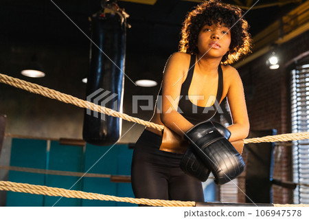 Woman fighter girl power. African american woman fighter with boxing gloves standing on boxing ring leaning on ropes waiting and resting. Strong powerful girl. Strength fit body workout training 106947578