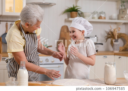 Happy family in kitchen. Grandmother and granddaughter child cook in kitchen together. Grandma teaching kid girl knead dough bake cookies. Household teamwork helping family generations concept 106947585