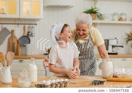 Happy family in kitchen. Grandmother and granddaughter child cook in kitchen together. Grandma teaching kid girl knead dough bake cookies. Household teamwork helping family generations concept Happy family in kitchen. Grandmother and granddaughter child cook in kitchen together. Grandma teaching kid girl knead dough bake cookies. Household teamwork helping family generations concept 106947588