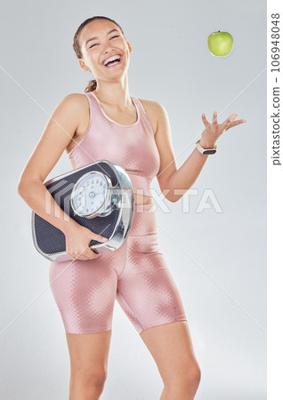 Happy woman, diet and scale to lose weight with apple, fitness goals or progress in studio. Portrait, laughing or female sports model throw fruit for healthy nutrition, weightloss results or wellness Happy woman, diet and scale to lose weight with apple, fitness goals or progress in studio. Portrait, laughing or female sports model throw fruit for healthy nutrition, weightloss results or wellness 106948048