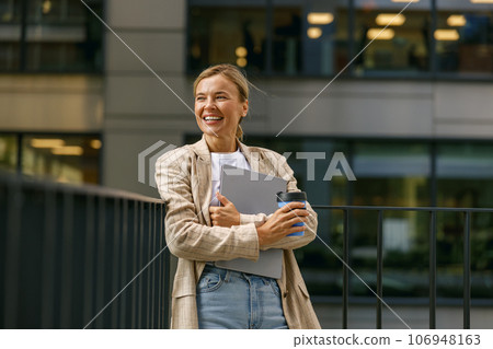 Business woman standing with laptop and coffee on office terrace during break time and looks away Business woman standing with laptop and coffee on office terrace during break time and looks away 106948163