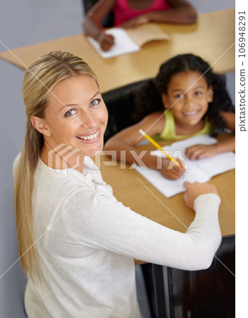 Kid, teacher and portrait in classroom studying school work for an education with support. Woman, smile and desk with child to help with reading or learning for children to work at school building. 106948291