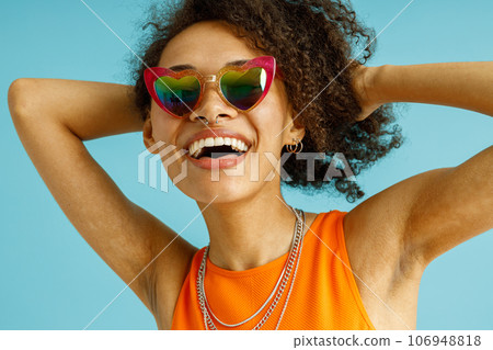 Close up of Smiling woman in heart-shaped glasses touching hair standing on blue studio background 106948818