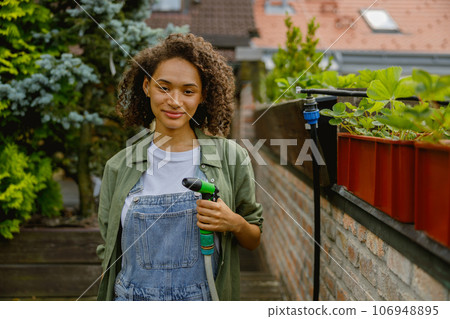 Woman gardener holding a hosepipe for watering plants in garden center and looks camera Woman gardener holding a hosepipe for watering plants in garden center and looks camera 106948895