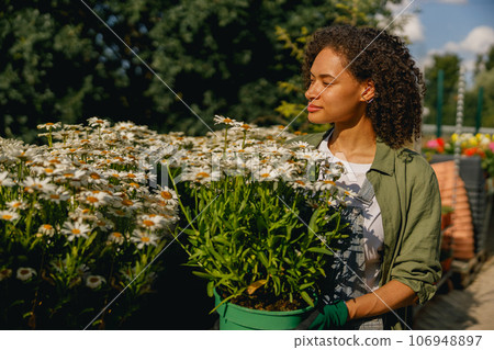 Pretty woman gardener holding flower pot while standing on greenhouse yard background 106948897