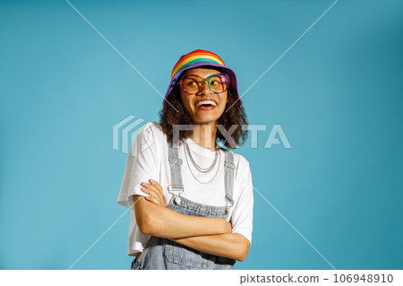 Smiling woman in rainbow cap with crossed hands looks away on blue studio background 106948910