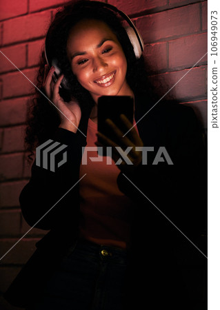 Woman, phone and headphones for city or night streaming, listening to club music or party in neon light. Happy young person on mobile, social media and audio for techno, rave or network on brick wall 106949073