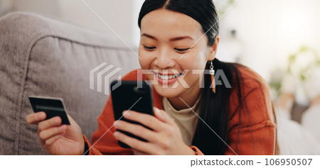 Asian woman, credit card and phone relaxing on her sofa online shopping, ecommerce and fintech easy payment. Young person in China typing bank information on cellphone for finance transaction at home 106950507