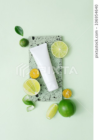 A white plastic tube with facial moisturizer cream or facial cleanser decorated with slices of lime and kumquat on gray brick podium on pastel background 106954640