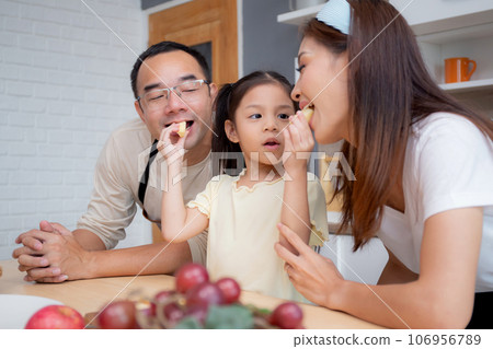 Happy family with father, mother and daughter in kitchen while daughter feed apple fruit. 106956789