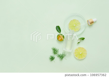 Glass petri dish containing lime slice with a dropper and bottle leaning on. Green leaves, kumquat and lime slices featured. Empty area in the left side for text adding 106957362