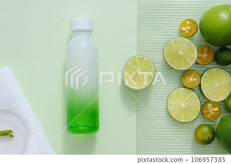 Unlabeled bottle in gradient green color displayed with many kumquat and lime round slices. White towel with a petri dish placed on. Template for branding mockup 106957385