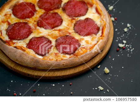 Hot tasty traditional italian pizza with salami, meat, cheese, tomatoes greens on a dark background Hot tasty traditional italian pizza with salami, meat, cheese, tomatoes greens on a dark background 106957580