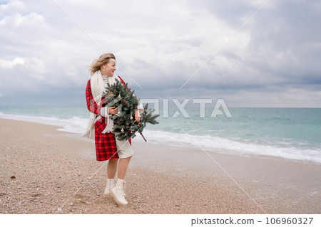 Blond woman holding Christmas tree by the sea. Christmas portrait of a happy woman walking along the beach and holding a Christmas tree in her hands. Dressed in a red coat, white dress. Blond woman holding Christmas tree by the sea. Christmas portrait of a happy woman walking along the beach and holding a Christmas tree in her hands. Dressed in a red coat, white dress. 106960327