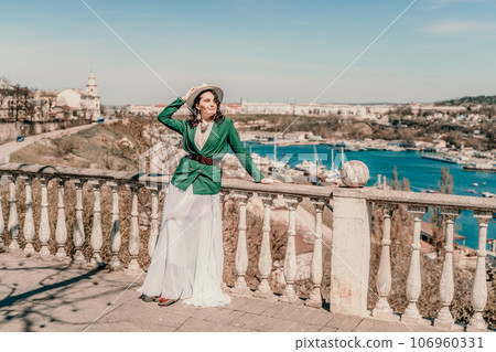 Woman walks around the city, lifestyle. Happy woman in a green jacket, white skirt and hat is sitting on a white fence with balusters overlooking the sea bay and the city. Woman walks around the city, lifestyle. Happy woman in a green jacket, white skirt and hat is sitting on a white fence with balusters overlooking the sea bay and the city. 106960331