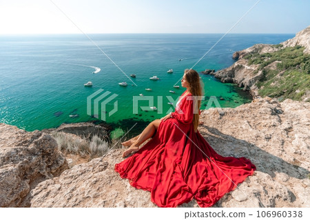 Woman red dress sea. Happy woman in a red dress and white bikini sitting on a rocky outcrop, gazing out at the sea with boats and yachts in the background. 106960338