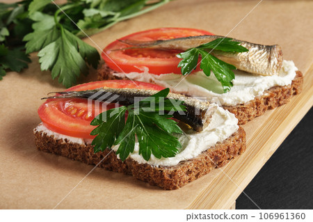 Sandwiches with sprats, tomatoes and parsley on wooden serving tray on black stone background. 106961360