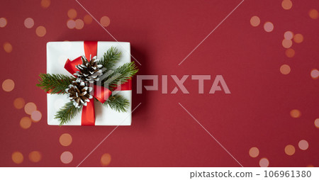 Banner with a white gift box with a red ribbon and fir twigs and cones on red background. Space for text. Banner with a white gift box with a red ribbon and fir twigs and cones on red background. Space for text. 106961380