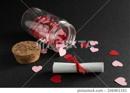 Small paper scroll tied with red rope. In background is small glass bottle with confetti hearts. Message in bottle. Valentines Gift 106961381