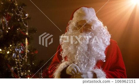 Santa Claus hangs a New Year's toy on the Christmas tree. A man dressed as Santa Claus looks stern. Santa is on camera and looks sternly. Santa Claus and Christmas tree. 106962151