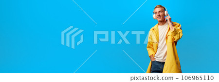 Banner. Portrait of young handsome smiling guy, student in bright fashion outfit and listening misic in headphones isolated on blue background. 106965110