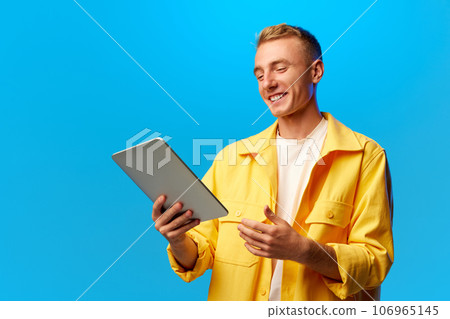 Portrait of young handsome smiling guy, student dressed in bright fashion outfit holding tablet isolated on blue background. 106965145
