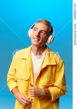 Portrait of young handsome guy, student in bright fashion outfit and headphones looking away isolated on blue background blue background. Portrait of young handsome guy, student in bright fashion outfit and headphones looking away isolated on blue background blue background. 106965150