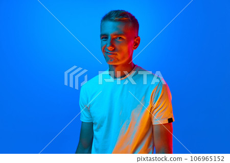 Portrait of young grimacing man, student in white t-shirt looking at camera isolated on blue background in red neon light, filter. Portrait of young grimacing man, student in white t-shirt looking at camera isolated on blue background in red neon light, filter. 106965152