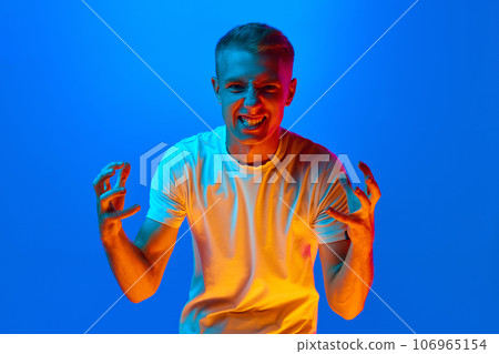 Portrait of young agressive, angry, irritated man, student in white t-shirt looking at camera isolated on blue background in red neon light, filter. Portrait of young agressive, angry, irritated man, student in white t-shirt looking at camera isolated on blue background in red neon light, filter. 106965154