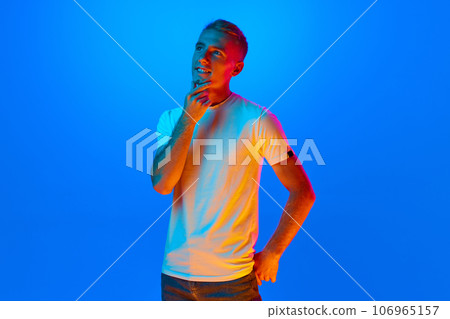Portrait of young man, student on blue studio background in neon light, filter. Handsome, dreaming male model in casual style. Portrait of young man, student on blue studio background in neon light, filter. Handsome, dreaming male model in casual style. 106965157