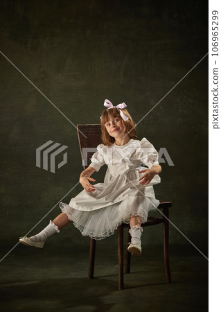 Portrait of little girl, child looks like dall, marionette in white dress sitting on chair against vintage background. Fairy princess or queen. Heroine of story tale. 106965299