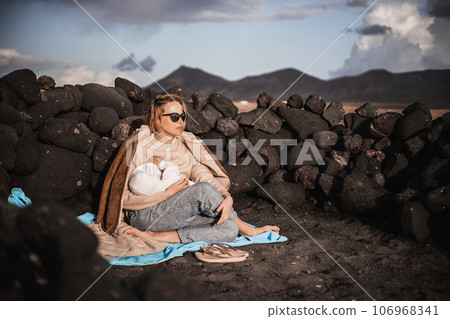 Mother breast feeding his infant baby boy son on black sandy volcanic beach of Janubio on Lanzarote island, Spain, enjoing dramatic volcanic landscape on windy overcast day. Travel with kids concept. 106968341