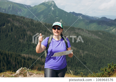 Stylish sport woman holding paper map, wearing backpack and hat looking at mountain view while relaxing in nature. Travel and wanderlust concept. Amazing chill moment. Redhead 30s girl giving thumb up Stylish sport woman holding paper map, wearing backpack and hat looking at mountain view while relaxing in nature. Travel and wanderlust concept. Amazing chill moment. Redhead 30s girl giving thumb up 106968576