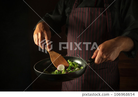 Cooking fresh vegetables in the restaurant kitchen. A frying pan with vegetables and a spatula in the hands of the chef. Vegetable diet concept on black background 106968654