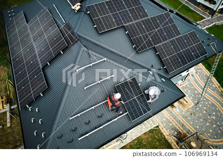 Engineers building photovoltaic solar module station on roof of house. Men electricians in helmets installing solar panel system outdoors. Concept of alternative and renewable energy. Aerial view. Engineers building photovoltaic solar module station on roof of house. Men electricians in helmets installing solar panel system outdoors. Concept of alternative and renewable energy. Aerial view. 106969134