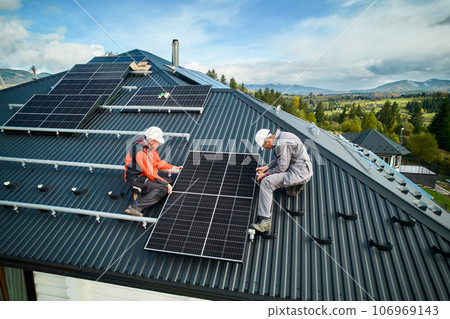 Roofers building photovoltaic solar module station on roof of house. Men electricians in helmets installing solar panel system outdoors. Concept of alternative and renewable energy. Aerial view. Roofers building photovoltaic solar module station on roof of house. Men electricians in helmets installing solar panel system outdoors. Concept of alternative and renewable energy. Aerial view. 106969143
