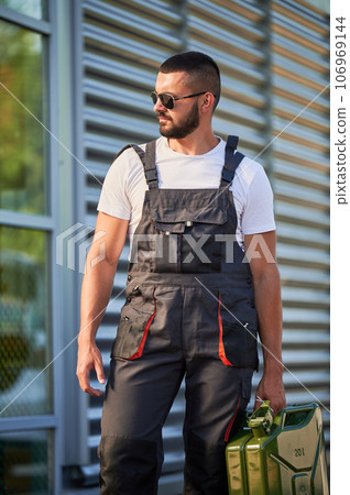Strong gas station worker holding canister full of fuel. Male operator caring heavy cistern with gasoline. Confident man in sunglasses and overalls with canister looking away. Strong gas station worker holding canister full of fuel. Male operator caring heavy cistern with gasoline. Confident man in sunglasses and overalls with canister looking away. 106969144