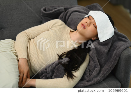 Sick young woman lying on couch with wet towel on her forehead for reduce high fever. Illness and health care concept Sick young woman lying on couch with wet towel on her forehead for reduce high fever. Illness and health care concept 106969601
